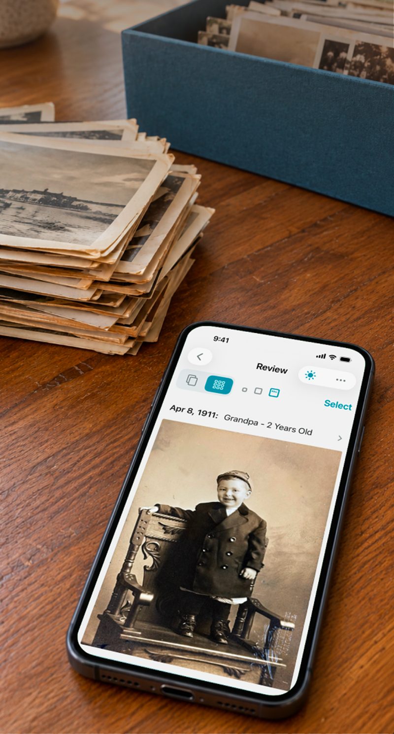 An iPhone on a table beside a stack of old prints, showing a scanned 1911 portrait captioned “Grandpa – 2 Years Old”